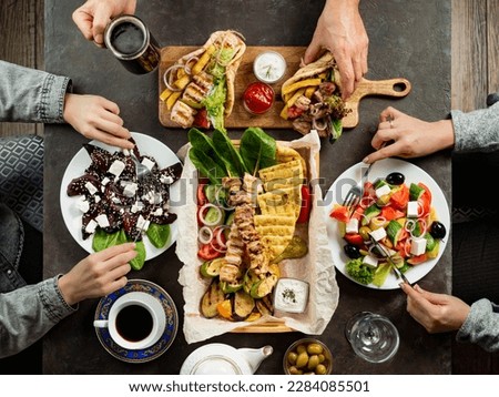 People eating Greek food. Greek cuisine assortment on dark tabletop, top down view. Hands with gyros, greek salad, souvlaki - selected of greek cuisine