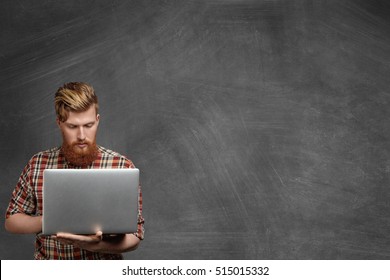 People and education concept. Hipster redhead student with fuzzy beard working on diploma project using laptop computer, standing at blank chalkboard in classroom, looking serious and concentrated