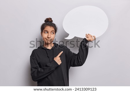 People emotions concept. Indoor shot of young fit glad Iranian girl standing in centre isolated on white background wearing black hoodie looking pointing and holding speech bubble for promotion