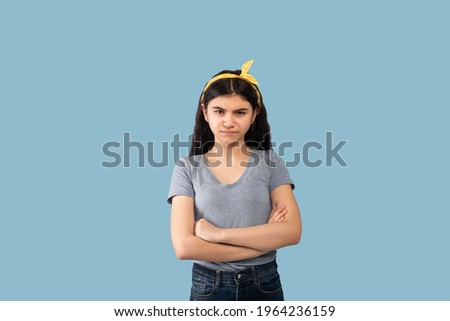 People emotions concept. Portrait of offended Indian teen girl posing with folded arms, looking at camera on blue studio background. Angry adolescent holding hands crossed, feeling dissatisfied