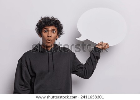 People emotions concept. Studio shot of young surprised Hindu man as if seeing something unexpected or unusual wearing black hoodie holding white speech bubble with blank space for promotion