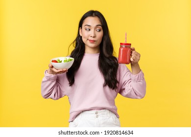 People emotions, healthy lifestyle and food concept. Curious cute asian girl looking at smoothie, holding salad and drink, making choice what eat for breakfast, standing yellow background