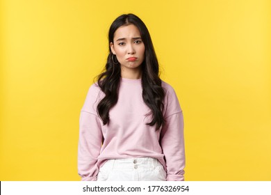 People emotions, lifestyle and fashion concept. Depressed and sad, gloomy korean girl pouting, looking down in dumps, feeling upset and displeased, standing yellow background