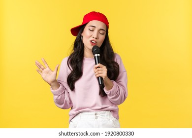 People Emotions, Lifestyle Leisure And Beauty Concept. Cool And Beautiful Asian Girl In Red Cap, Close Eyes And Singing In Microphone, Performing On Stage, Standing Yellow Background