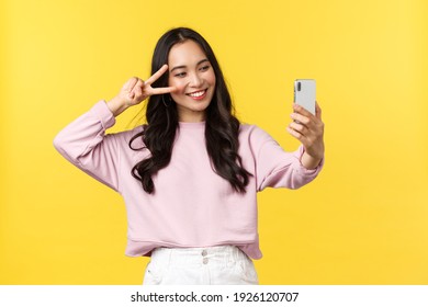 People emotions, lifestyle leisure and beauty concept. Cheerful asian girl over yellow background taking selfie on mobile phone, use photo filters app and show peace sign near face and smiling