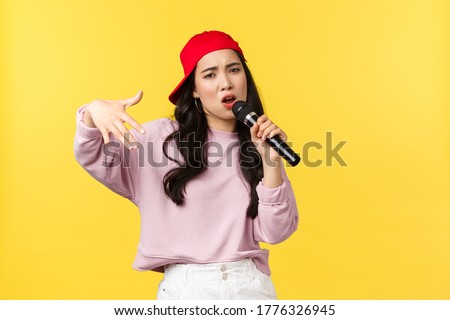 People emotions, lifestyle leisure and beauty concept. Stylish and cool young girl rapper in red cap, singing song and gesturing, performing with microphone, standing yellow background