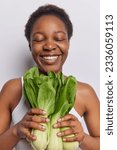 People emotions and natural beauty. Studio vertical photo of young cheerful satisfied smiling broadly African girl standing in centre on white background holding bunch of bok choy. Healthy lifestyle
