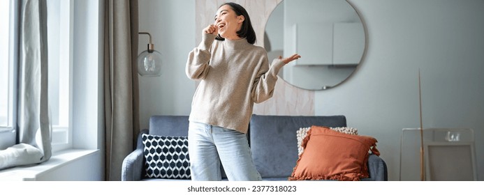 People and emotions. Portrait of asian girl standing in living room, singing in shadow microphone, dancing and having fun, feeling joy. - Powered by Shutterstock