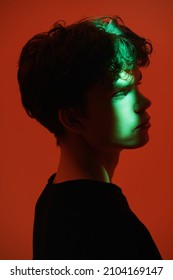 People, Emotions. A Portrait Of A Handsome Dark-haired Guy With A  Beam Of Light On His Face Looking Sadly And Pensively Away. Sideview Shot On A Red Background. People And Emotions. 