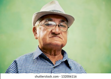 People And Emotions, Portrait Of Serious Senior Hispanic Man With Glasses And Hat Looking At Camera Against Green Background