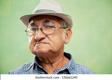 People And Emotions, Portrait Of Serious Senior Hispanic Man With Glasses And Hat Looking At Camera Against Green Background