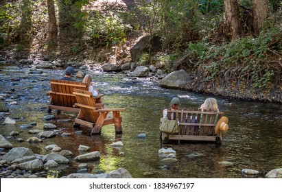 People Enjoy The Afternoon Sitting In Wooden Chairs Placed In The Big Sur River, In California. 