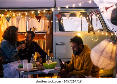 People enjoying camping lifestyle and van life travel wanderlust - caucasian men and woman have fun and dinner outside a vintage van - evening time with lights and romance