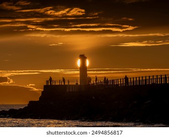 People enjoying a scenic walk on a pier near a tall lighthouse with the sun setting in the background - Powered by Shutterstock