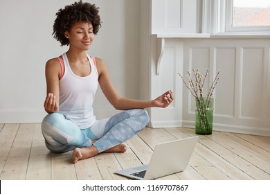 People, Ethnicity, Yoga And Meditation Concept. Beautiful Pleased Delighted Dark Skinned Relaxed Student Sits Crossed Legs On Floor Near Opened Laptop, Tries To Have Break After Work On Diploma Paper