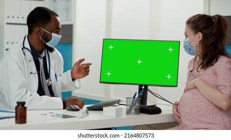 People With Face Mask Using Computer With Greenscreen At Pregnancy Checkup Visit. Woman And Medic Analyzing Blank Copyspace Background With Isolated Mockup Template And Chroma Key. Tripod Shot.