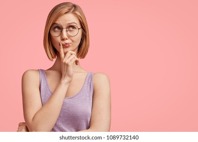 People, facial expressions and thoughts. Attractive young female student looks pensively upwards, wears spectacles, poses against pink background with blank copy space for your advertisement