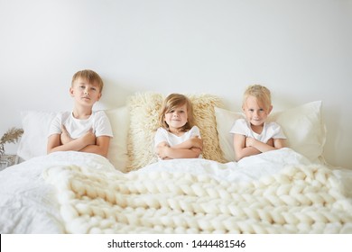 People, Family And Childhood Concept. Three Kids Sitting Next To Each Other On Large White Bed With Arms Folded, Watching Cartoons On Weekend Morning. Two Brothers And Sister Playing At Home