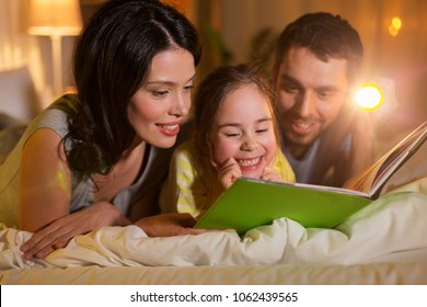 People And Family Concept - Happy Mother, Father And Little Daughter Reading Book With Torch Light In Bed At Night At Home