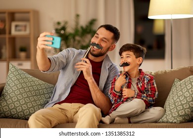 people, family and technology concept - happy father and little son with mustaches party props taking selfie by smartphone sitting on sofa at home in evening - Powered by Shutterstock