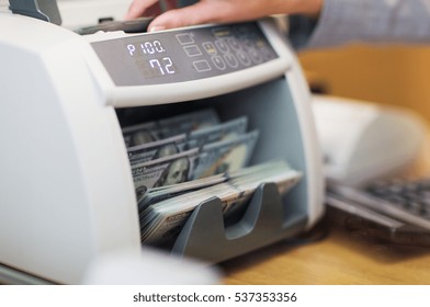 People, Finance And Cash Concept - Clerk Hand Counting Dollars With Electronic Money Counter Machine At Bank Office Or Currency Exchanger