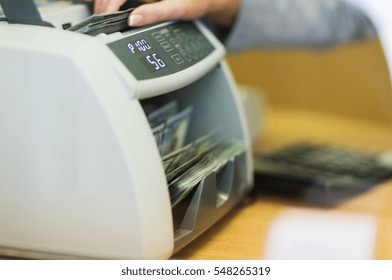 People, Finance And Cash Concept - Clerk Hand Counting Dollars With Electronic Money Counter Machine At Bank Office Or Currency Exchanger