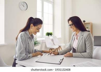 People And Finance. Smiling Bank Manager Sitting At Office Desk And Asking Client To Sign Loan Agreement. Happy Woman Meeting With Business Adviser And Talking About Contract Terms And Conditions