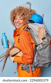 People free time and camping concept. Cheerful female tourist has hiking adventure going to make campfire and have picnic carries wood poses with rucksack full of equipment wears hat and jacket