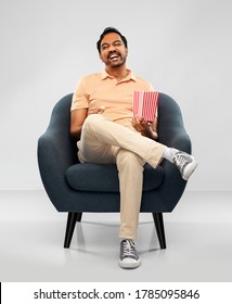 people and furniture concept - happy smiling young indian man eating popcorn sitting in chair over grey background - Shutterstock ID 1785095846