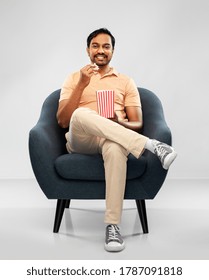 people and furniture concept - happy smiling young indian man eating popcorn sitting in chair over grey background - Shutterstock ID 1787091818