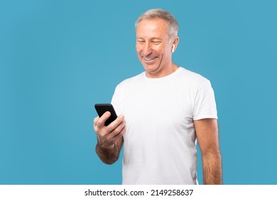 People And Gadgets. Portrait of happy smiling mature man using cell phone wearing wireless earbuds standing isolated over blue studio background. Adult male chatting online, listening to music - Powered by Shutterstock