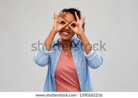 people, gesture and portrait concept - happy african american young woman looking through finger glasses over grey background
