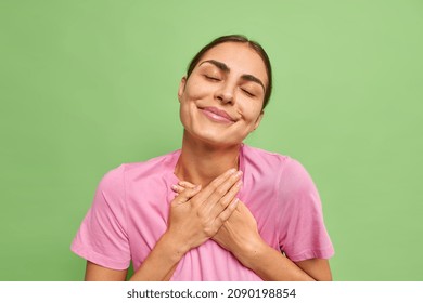 People And Gratitude Concept. Pleased Brunette Woman Says Thank You Holds Hands On Heart Feels Grateful For Gift Or Help Keeps Eyes Closed Dressed In Casual Pink T Shirt Isolated Over Green Background