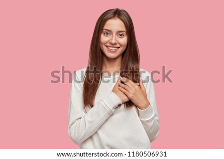 People, gratitude, positive feeling concept. Satisfied generous brunette woman with tender smile, keeps both palms on chest, wears casual sweater, stands against pink background, being kind hearted