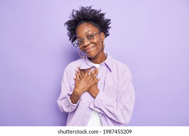 People gratitude and truthful feelings concept. Generous pretty dark skinned woman presses hands to chest makes thankful gesture being kind hearted wears spectacles shirt poses over purple wall