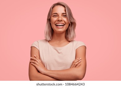 People, happiness and facial expressions concept. Pretty young woman with broad shining smile, keeps hands crossed, being in high spirit, wears braces on teeth, poses alone against pink background - Shutterstock ID 1084664243