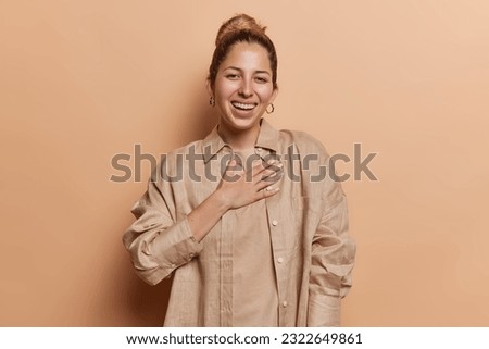 People and happiness. Sincere young European woman keeps hand on chest laughs gladfully expresses positive emotions smiles broadly dressed in shirt isolated over brown background being in good mood