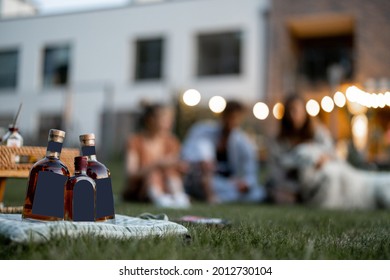 People Have Fun On The Picnic At Backyard In The Evening, Close-up On Bottles In Front With Blank Label To Copy Paste