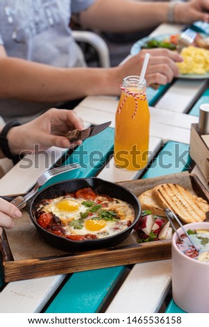 People are having breakfast on summer terrace: israeli shakshuka with tomatoes, salami and fried eggs, orange juice, omelette and light snacks