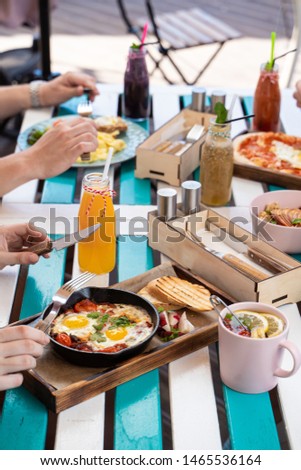 People are having breakfast on summer terrace: israeli shakshuka with tomatoes, salami and fried eggs, orange juice, omelette and light snacks
