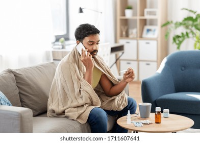 People, Health And Fever Concept - Sick Indian Man In Blanket With Thermometer Calling On Smartphone At Home