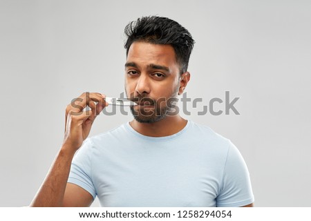 people, healthcare and fever concept - unhealthy indian man measuring oral temperature by thermometer over grey background