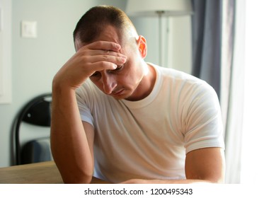 People, healthcare, stress and problem concept. Unhappy man suffering from headache at home. Copy space. A man with a headache sits in the dining room bowed head toward a wooden table.