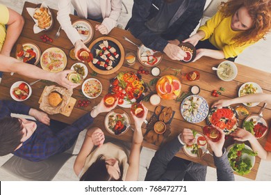 People hold glasses and eat healthy meals at party dinner table. Friends celebrate with organic food, ratatoille and corn barbecue on wooden table top view.