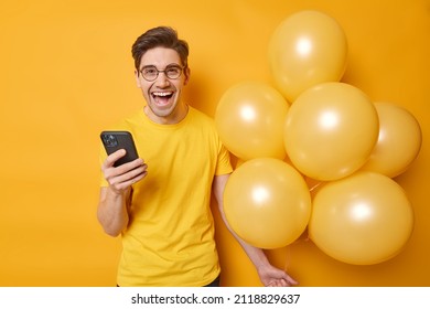 People holidays and celebration concept. Happy dark haired man uses mobile phone for sending text messages holds bunch of inflated balloons isolated over vivid yellow background ready for party