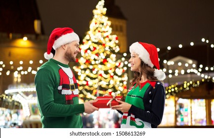 People And Holidays Concept - Happy Couple In Santa Hats And Ugly Sweaters With Gift Box Over Christmas Market Lights Background