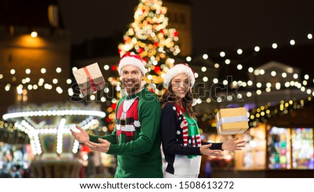 people and holidays concept - happy couple in santa hats and ugly sweaters with gifts over christmas market lights background