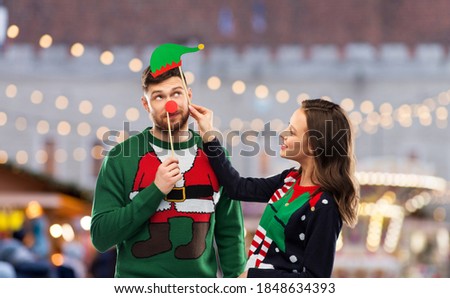 people and holidays concept - happy couple in ugly sweaters posing with party props over christmas market background