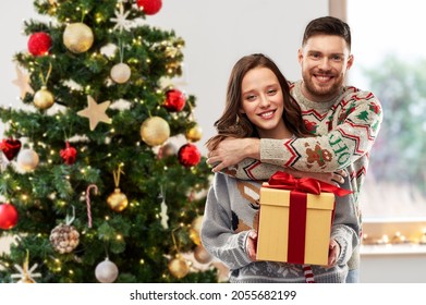 People And Holidays Concept - Portrait Of Happy Couple With Gift At Ugly Sweater Party Over Christmas Tree On Background