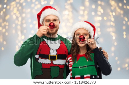 people and holidays concept - portrait of happy couple in santa hats making noses of red christmas balls at ugly sweater party over festive lights background